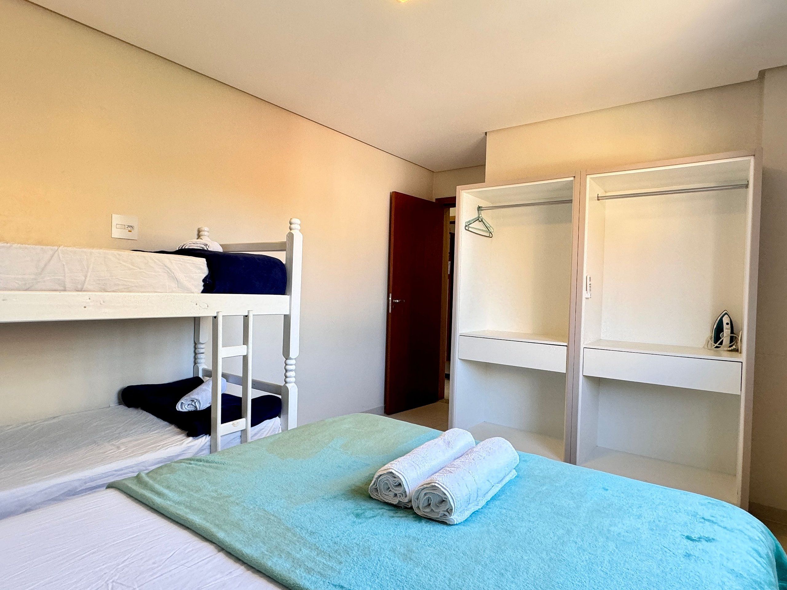 A dormitory room with bunk beds, a green bedspread, and a wardrobe with hangers.