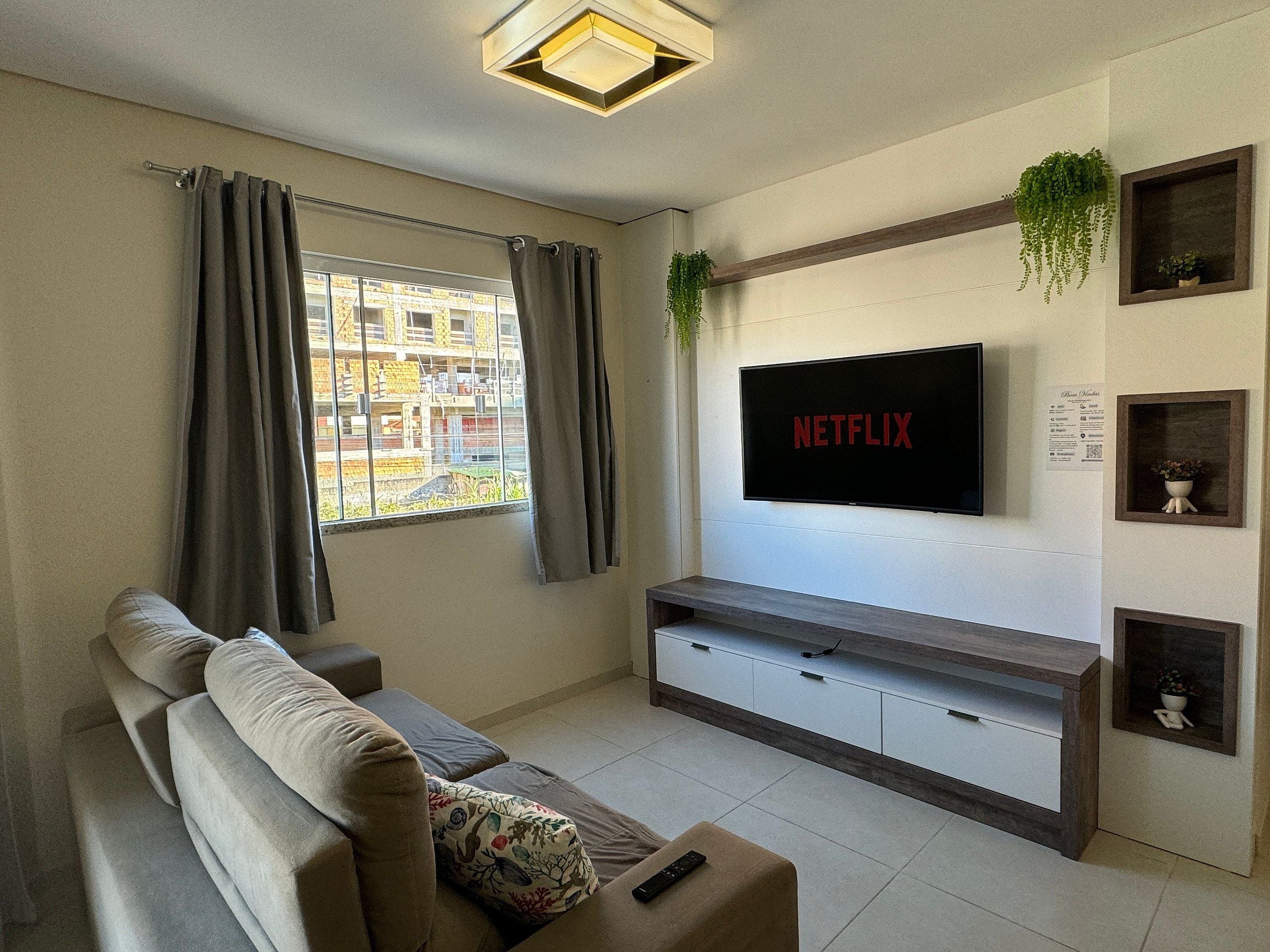 A living room with a large flat-screen TV mounted on the wall, a sofa, a coffee table, and a window with curtains.