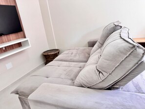 A modern living room with a grey sofa, a mounted flat-screen TV, and a small wooden side table. - Maria Julia (Bombinhas)