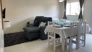 A dining area with a table set for four, a grey sofa with patterned cushions, and a ceiling fan. - Maria Julia (Bombinhas)