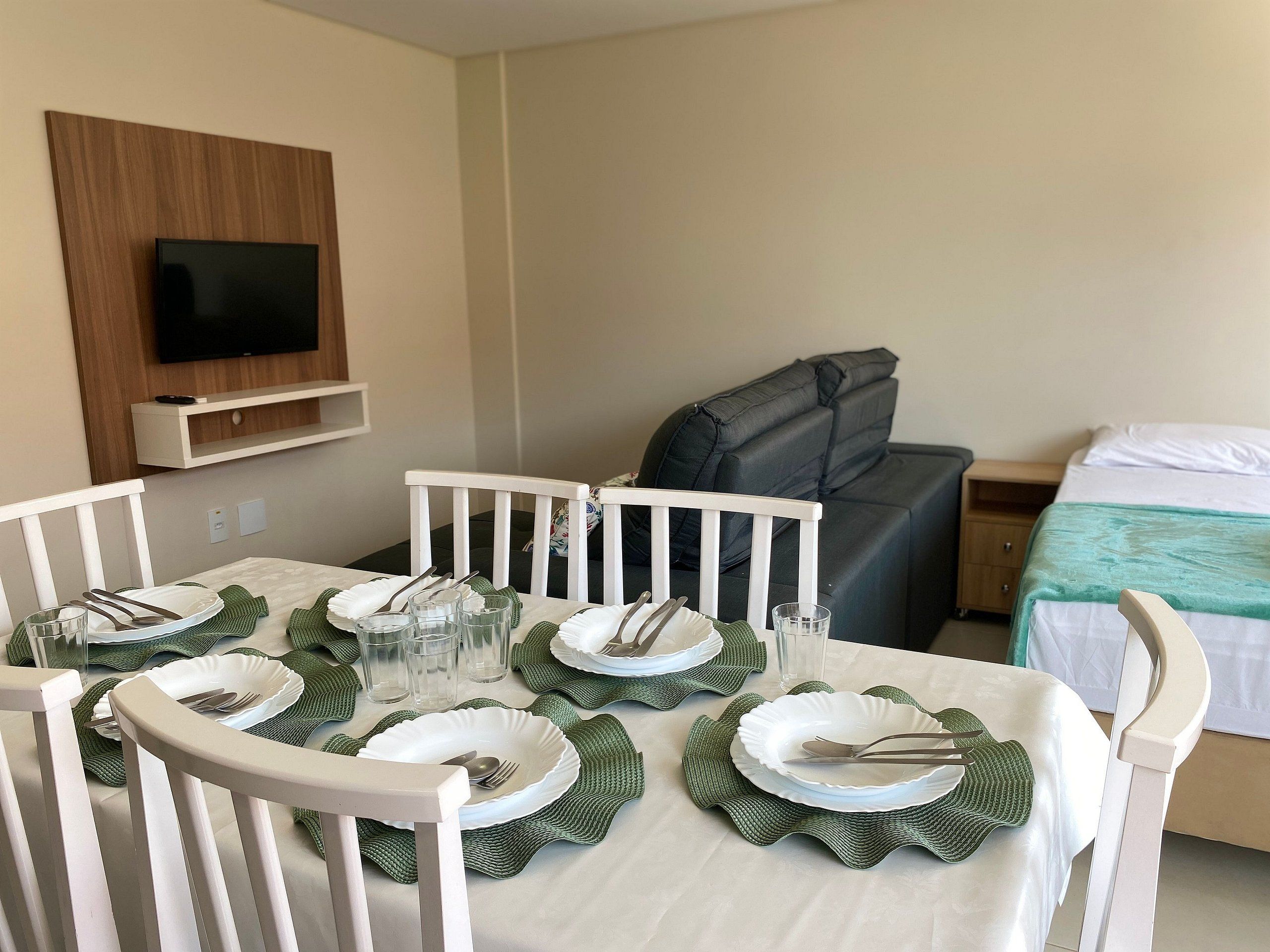 A neatly set dining table with white chairs, a television mounted on the wall, and a bed with a green blanket in a hotel room.