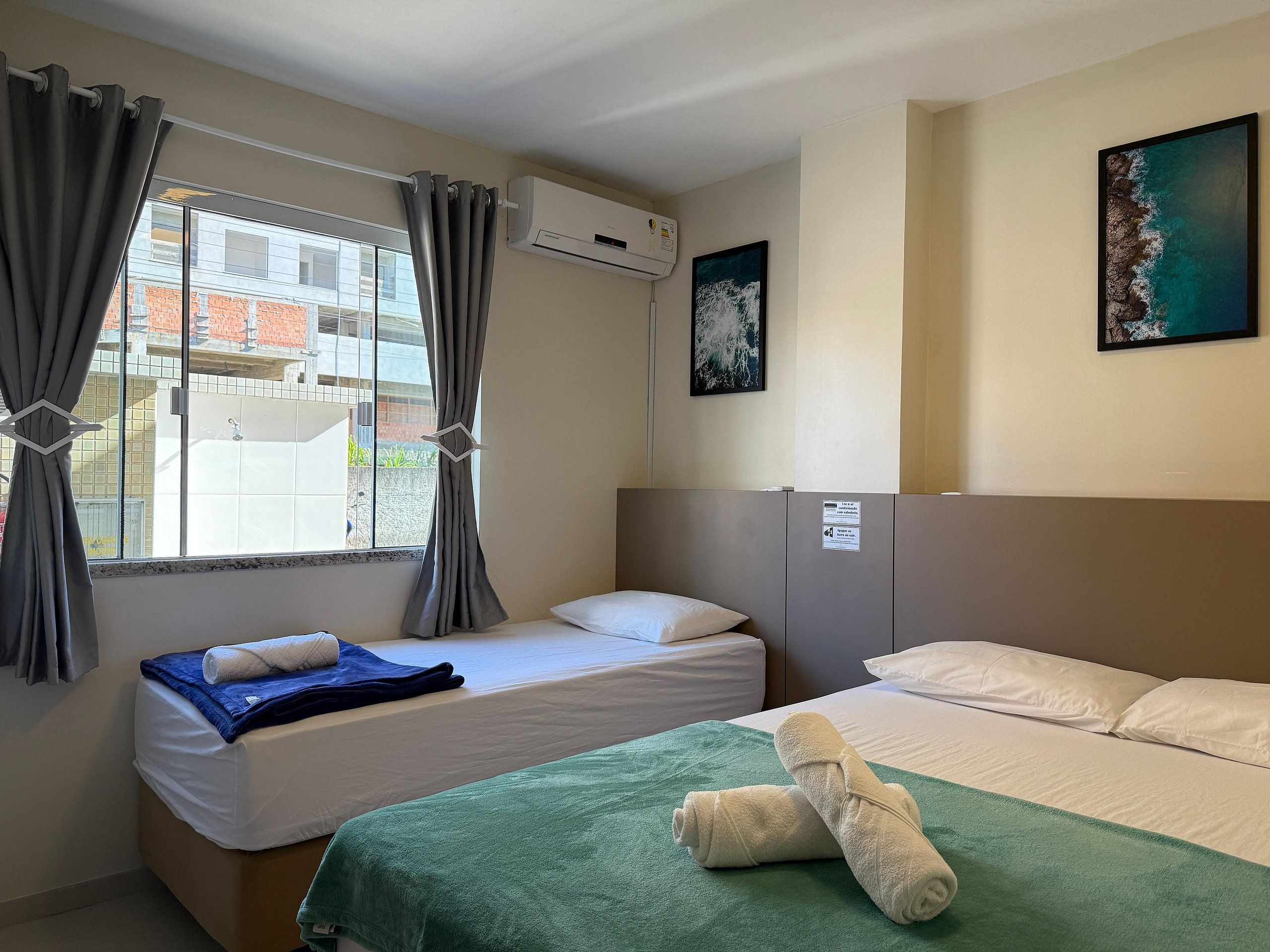 A hotel room with two beds, a window with curtains, an air conditioning unit, a painting on the wall, and a green towel on the bed.