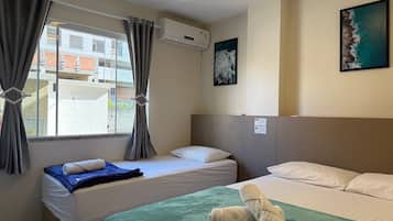 Quarto de hotel com duas camas, uma janela com cortinas, um ar condicionado, um quadro na parede e uma toalha verde sobre a cama.