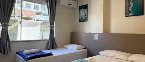 A hotel room with two beds, a window with curtains, an air conditioning unit, a painting on the wall, and a green towel on the bed.