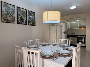 Un comedor con una mesa redonda dispuesta para seis personas, rodeada de sillas blancas. Hay cuadros con motivos de hojas tropicales en la pared, un lámpara colgante arriba y una cocina al fondo.