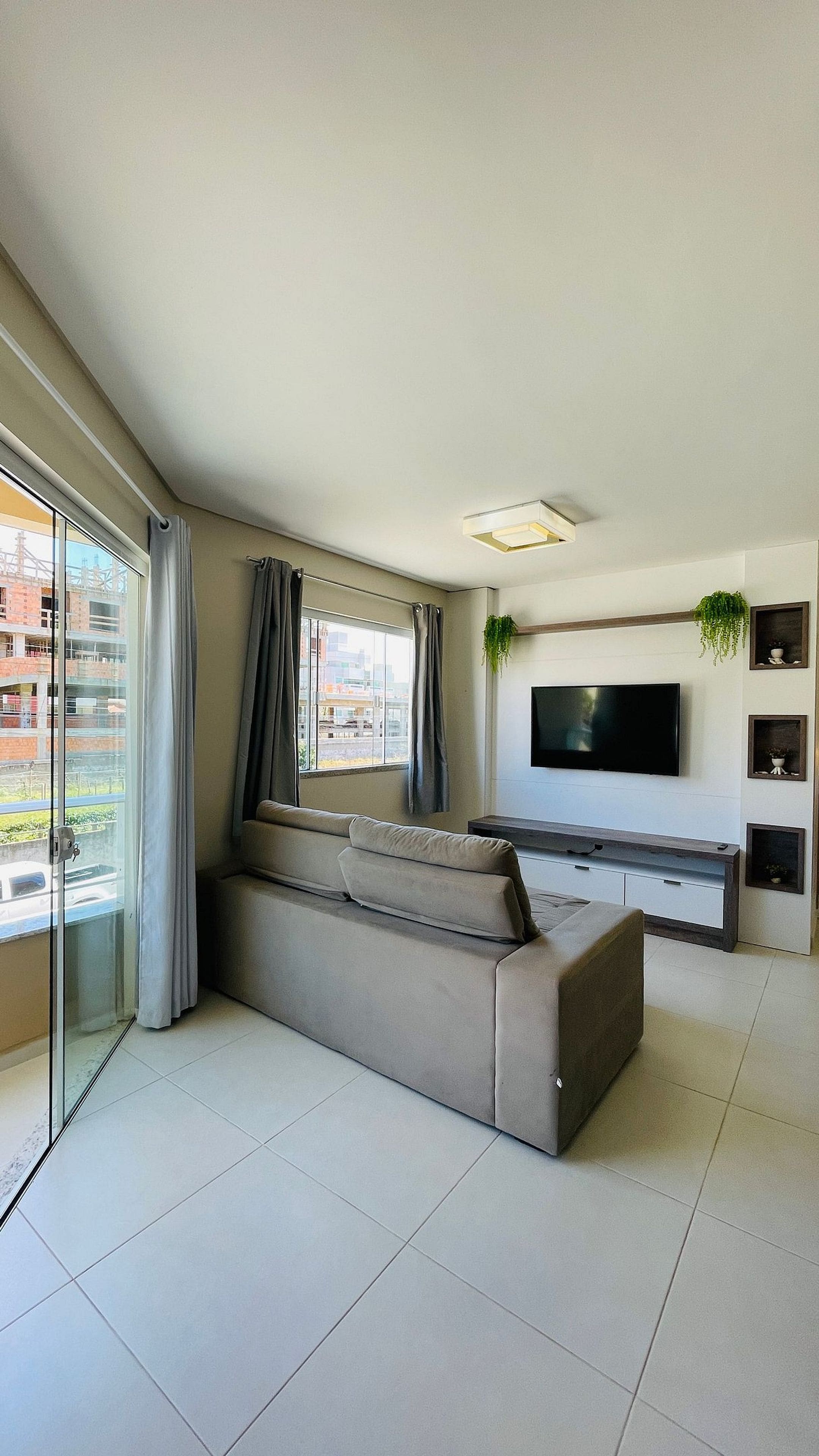 Un salón moderno con un sofá grande, un televisor de pantalla plana montado en la pared y un ventanal corredera que da a un balcón exterior.
