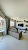 Uma sala de estar moderna com um sofá grande, uma TV de tela plana na parede e uma porta de vidro deslizante para uma varanda externa.