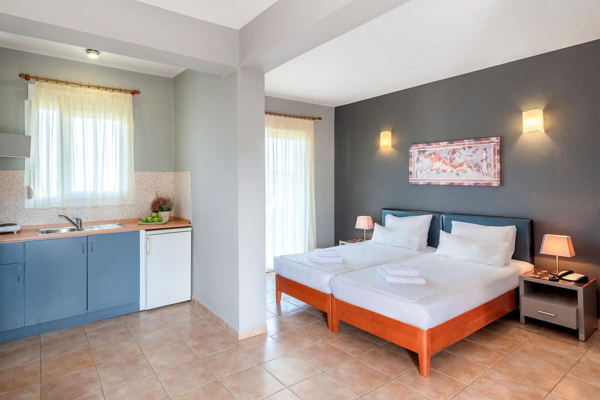 Una habitación de hotel con cama, mesitas de noche, un escritorio pequeño, ventana con cortinas, zona de cocina con fregadero y armarios, y una obra de arte colgada en la pared.