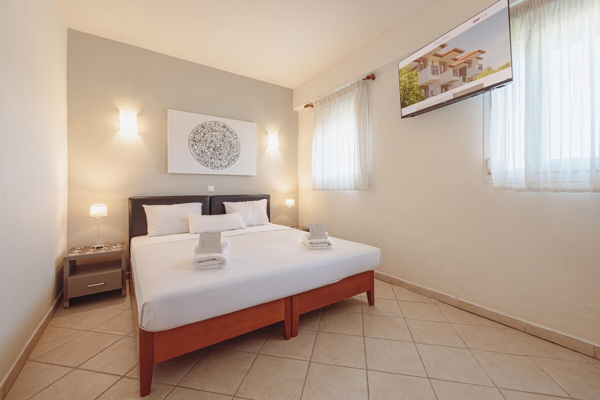 A neatly made bed with white linens and two pillows, a bedside table with a lamp, and a wall-mounted TV displaying a building image.