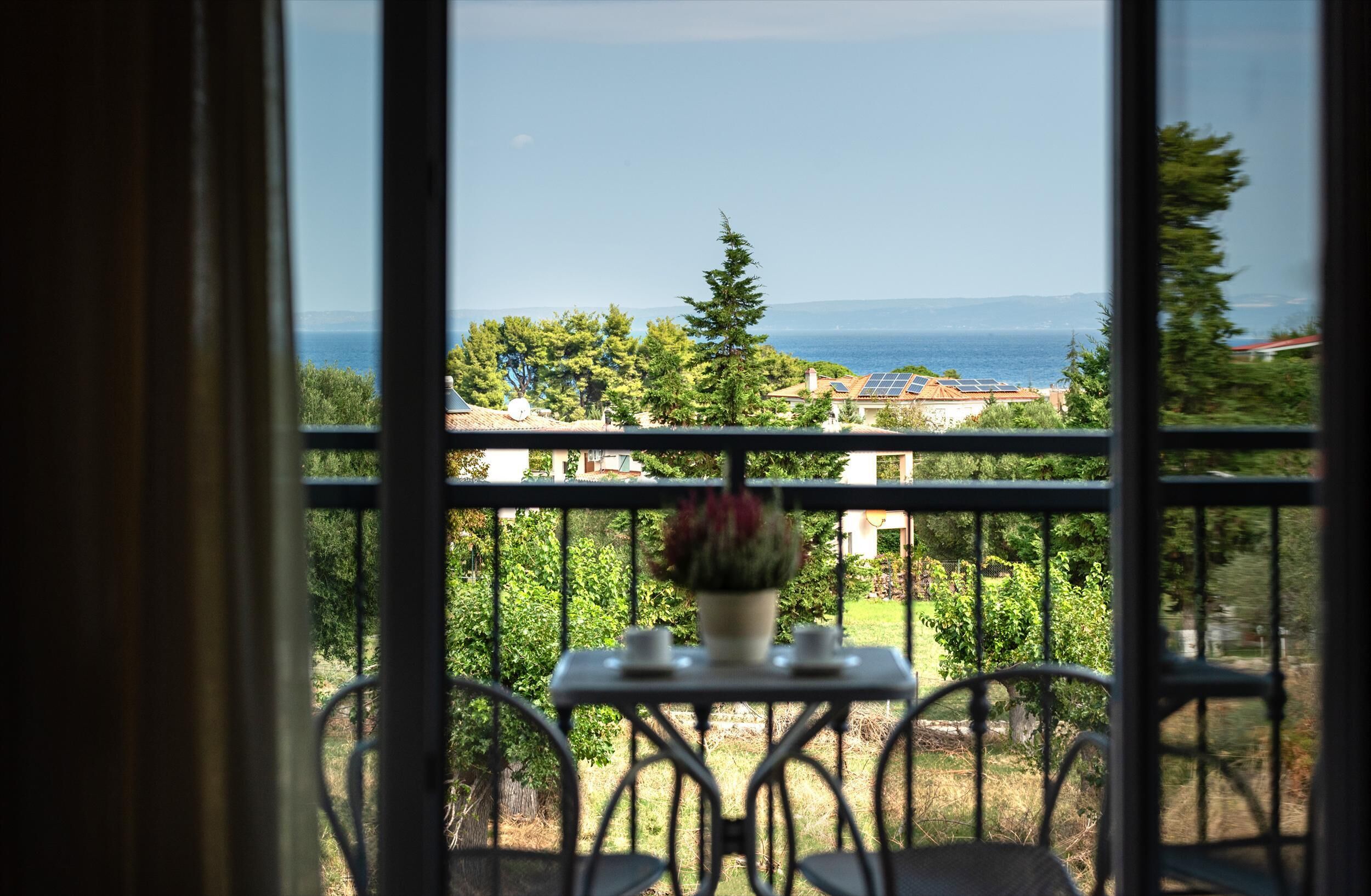 A balcony with a table and chairs, a potted plant, and a view of the sea and distant buildings.