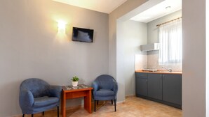 A small, modern kitchen with two blue chairs, a small table with a plant, and a flat-screen TV mounted on the wall.