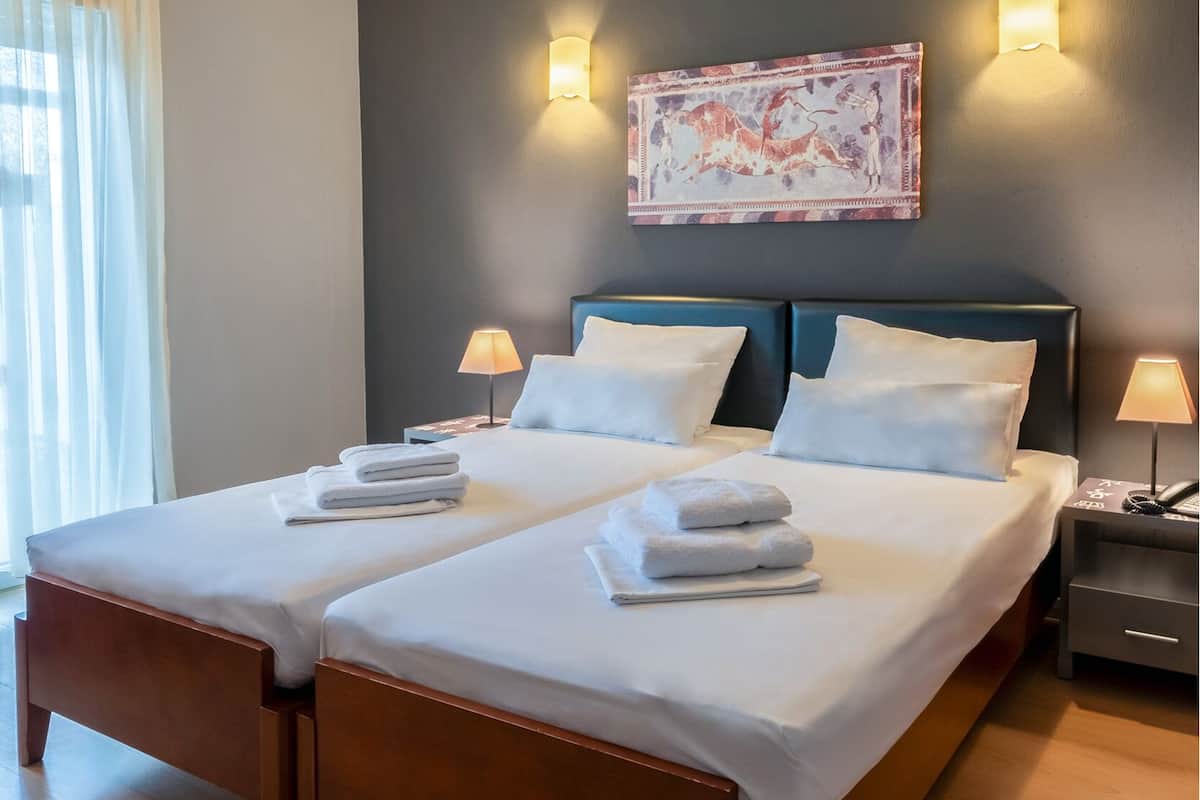 Una cama doble con ropa de cama y toallas blancas, dos mesitas de noche con lámparas, una obra de arte en la pared y suelo de madera.