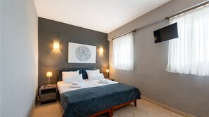 A neatly made bed with a dark blue bedspread, two bedside lamps, a flat-screen TV mounted on the wall, and a large framed artwork above the bed.