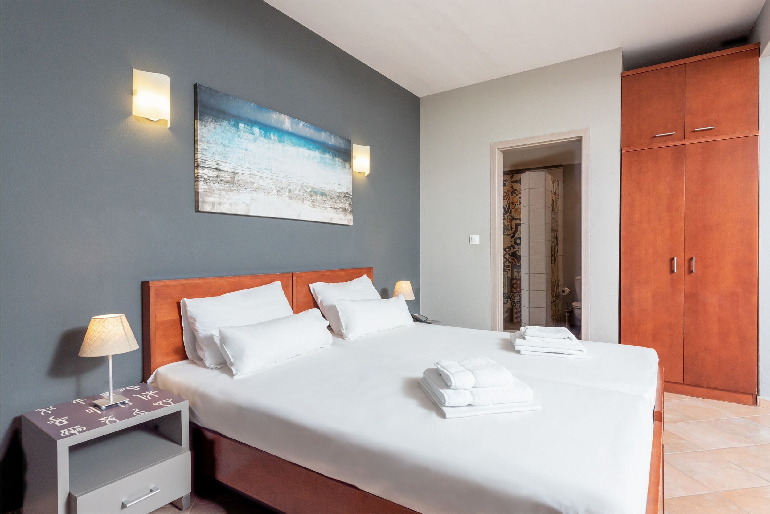 Een opgemaakt bed met witte lakens en handdoeken, een nachtkastje met een lamp, een schilderij aan de muur en een kledingkast.