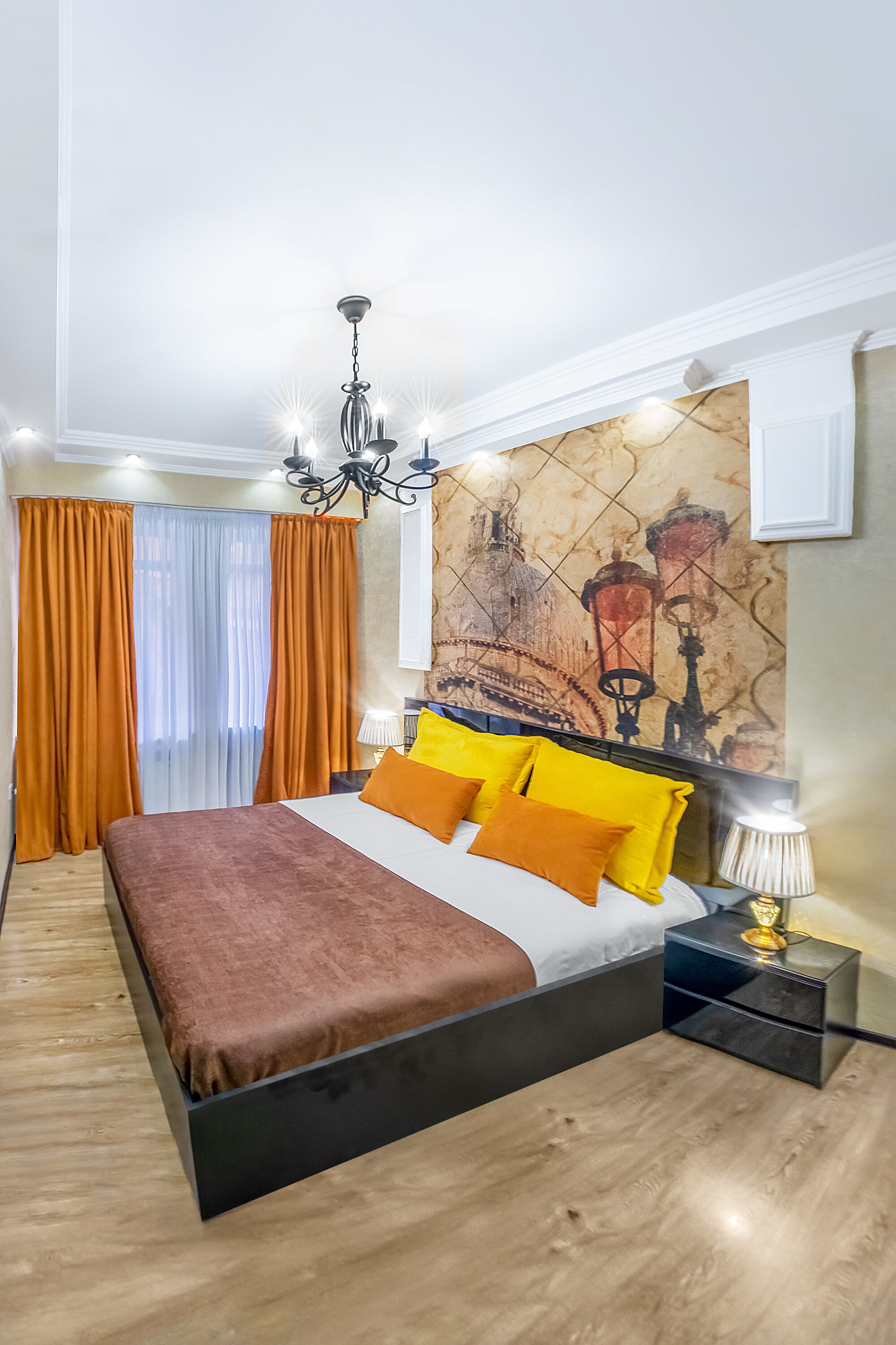 A modern bedroom with a large bed, orange curtains, a chandelier, and a cityscape painting on the wall.