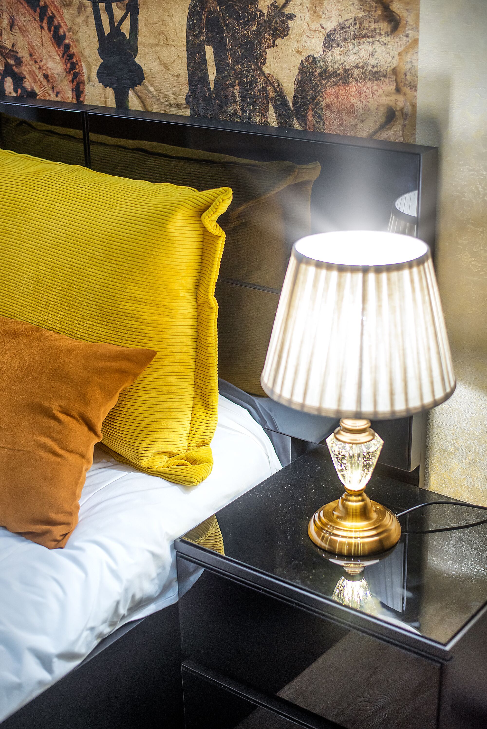 Een bed met gele en bruine kussens, een zwart nachtkastje en een lamp met gouden voet.
