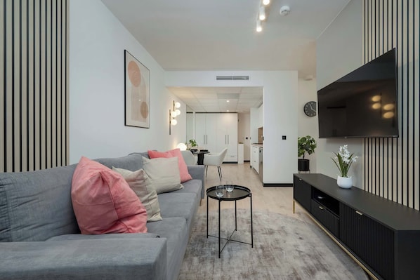 Un salon moderne avec un canapé gris, une petite table ronde, un meuble TV noir et une cuisine en arrière-plan.