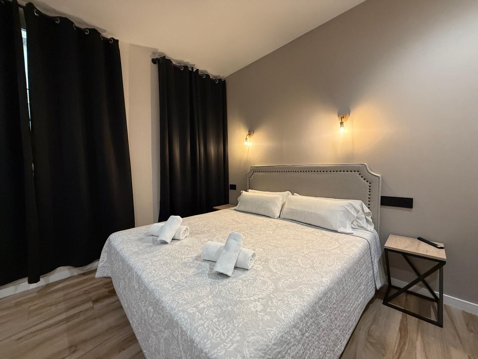 Una cama bien tendida con ropa blanca y toallas, una mesita de noche de madera y dos luces fijadas a la pared.