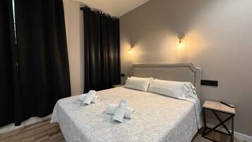 Una cama bien tendida con ropa blanca y toallas, una mesita de noche de madera y dos luces fijadas a la pared.