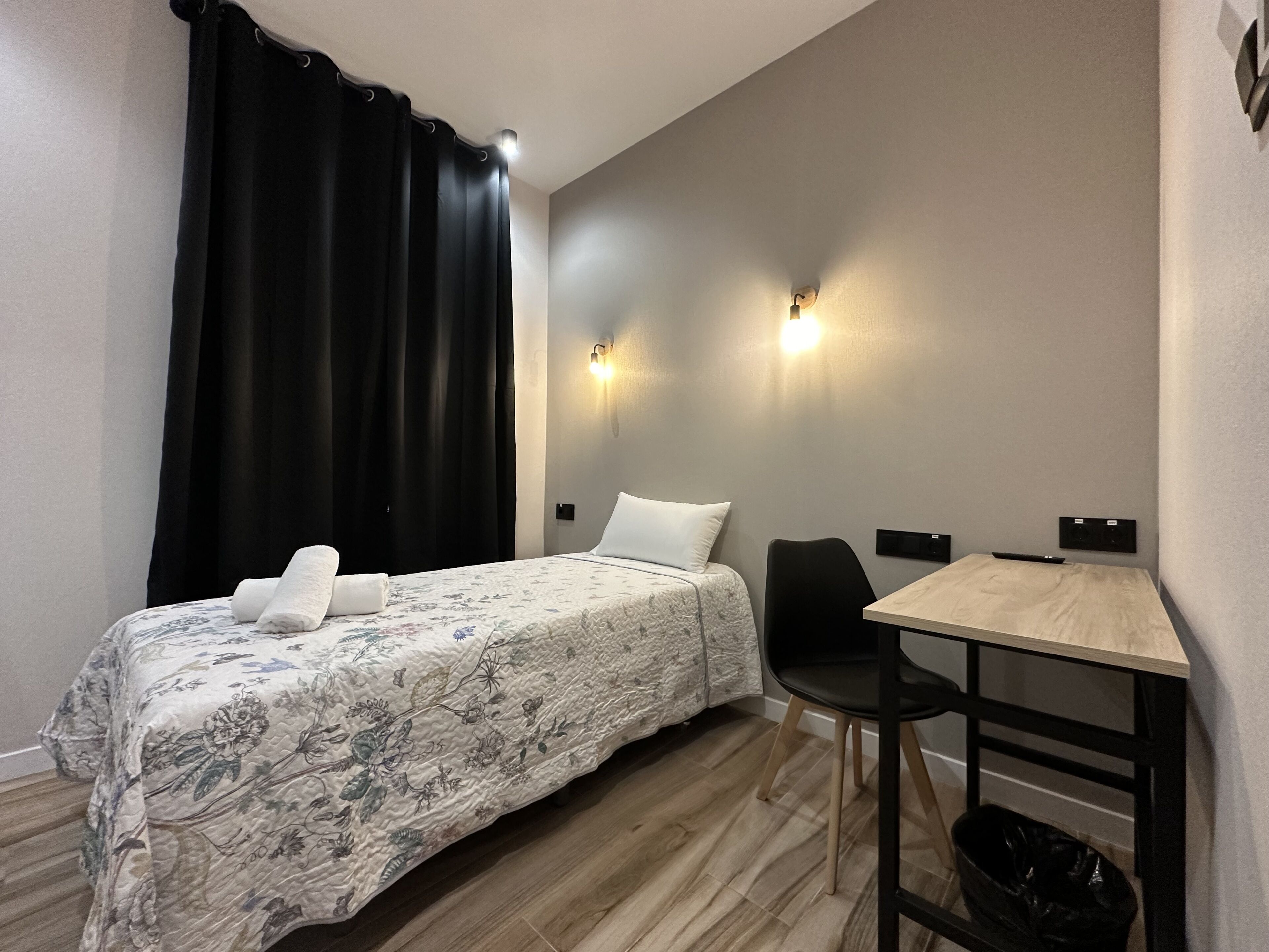 A hotel room with a single bed, a chair, a small table, and two wall-mounted lights.