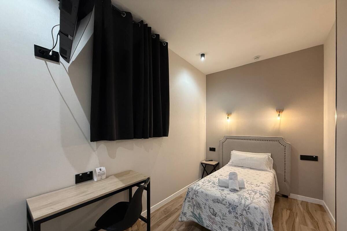 A bedroom with a bed, a desk, a chair, a computer, and two wall-mounted lights.