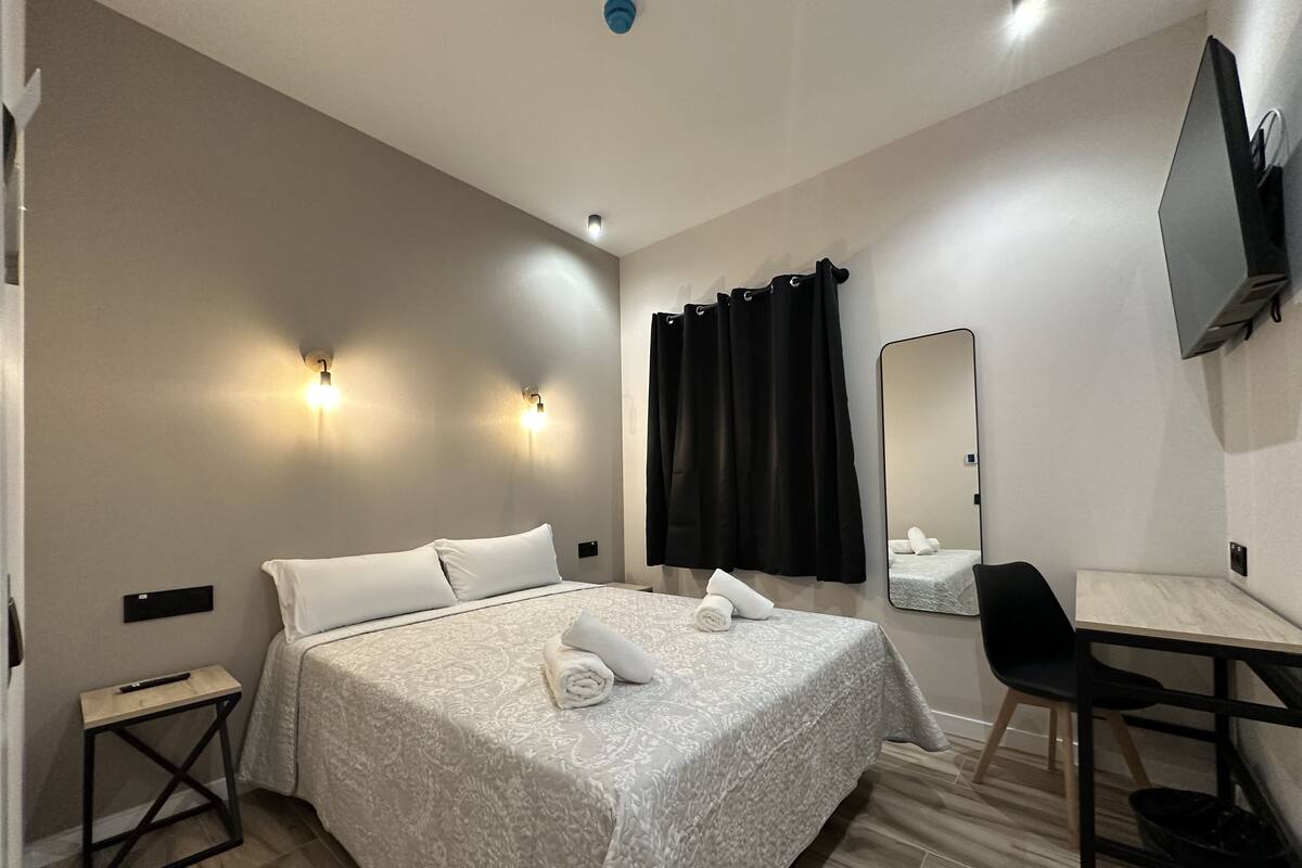 A hotel room with a bed, a desk, a chair, a mirror, and a TV.