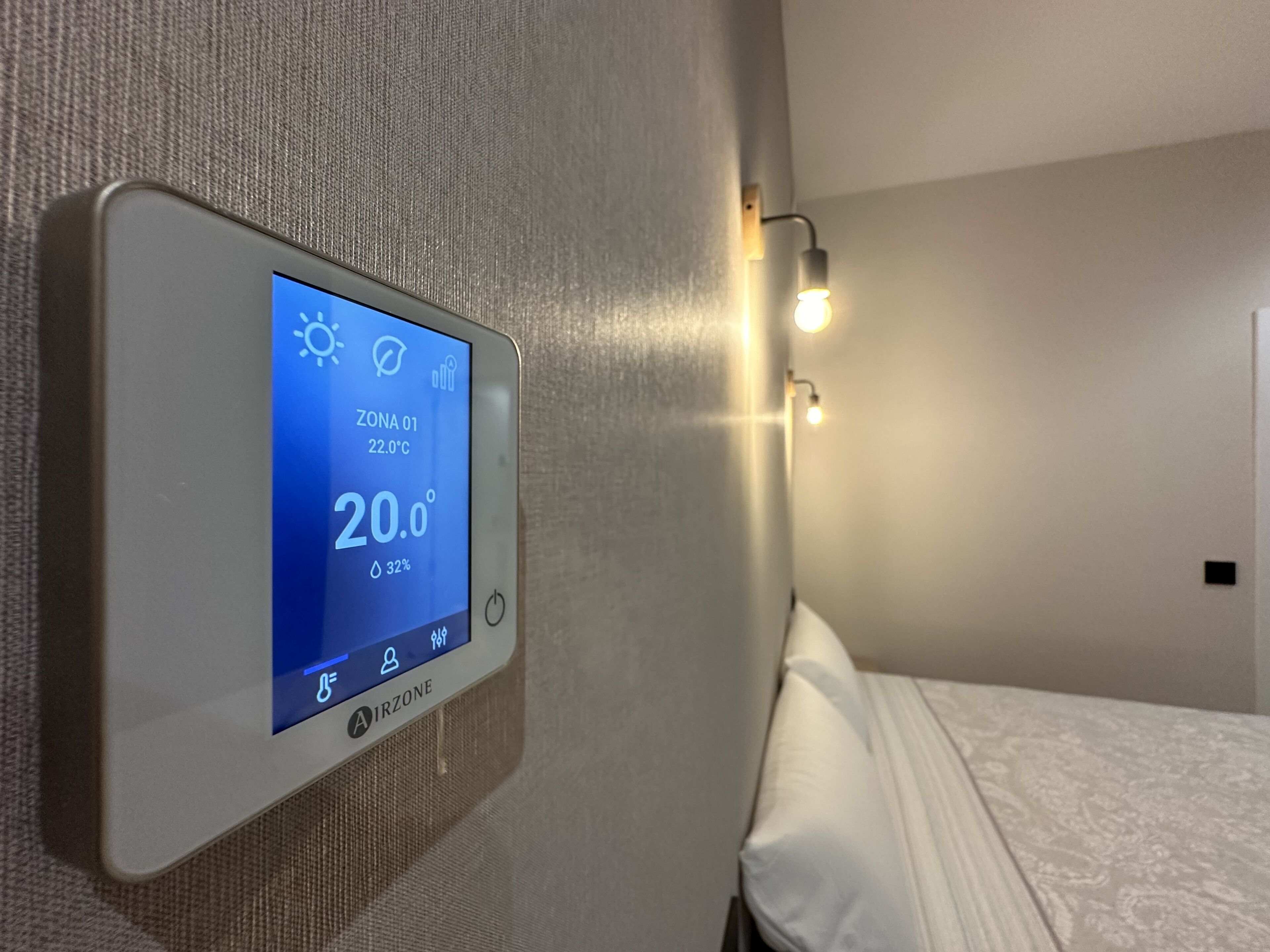 A thermostat displaying 20.0°C and 32% humidity in a room with a bed and a wall-mounted light fixture.