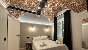 A modern bedroom with a brick ceiling, a large bed with a patterned bedspread, two bedside lamps, a small table, and a window with curtains. - CC Opera Suites (Madrid)