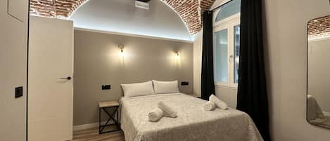 A modern bedroom with a brick ceiling, a large bed with a patterned bedspread, two bedside lamps, a small table, and a window with curtains.