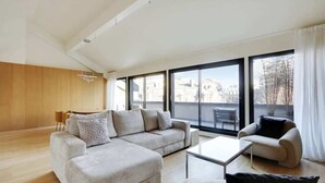 A spacious living room with a large sofa, a smaller sofa, and a coffee table. There is a wooden panel wall on one side and large windows on the other. - A Stunning 2-BR/2BA with Terraces and A/C (Paris)
