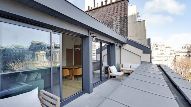 A modern building with large glass windows, a rooftop terrace with outdoor seating, and a view of the cityscape. - A Stunning 2-BR/2BA with Terraces and A/C (Paris)