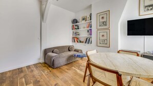 A modern living room with a grey sofa, a round wooden dining table, wicker chairs, a bookshelf with various books and framed art, and a flat-screen TV mounted on the wall. - A Nice 1-BR in Saint-Germain (Paris)
