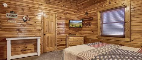 Une chambre de style cabane en bois, équipée d’un lit, d’une télévision, d’un bureau et d’une fenêtre avec des stores.