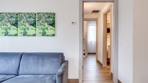 A modern living room with a grey sofa, a hallway leading to a bedroom, and a wall-mounted painting of trees. - Tenuta di Palù (Bardolino)