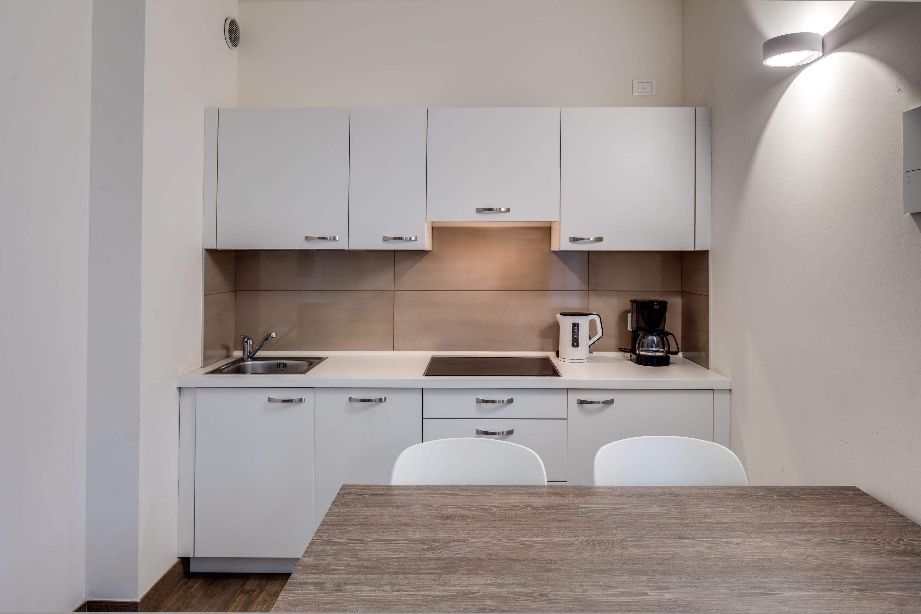 A modern kitchen with white cabinets, a sink, a coffee maker, and a wooden table.