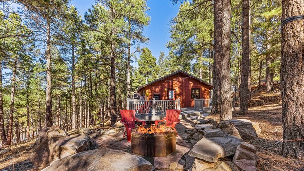 A cabin surrounded by tall pine trees, with a stone fire pit and red chairs in the foreground. - Chaco Trail | 2BR / 2BA | | Spend Spring in Flagstaff! (Flagstaff)