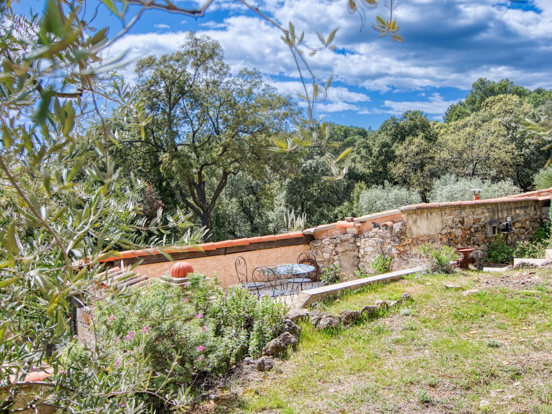 A rustic stone wall with a metal table and chairs, surrounded by greenery and trees.