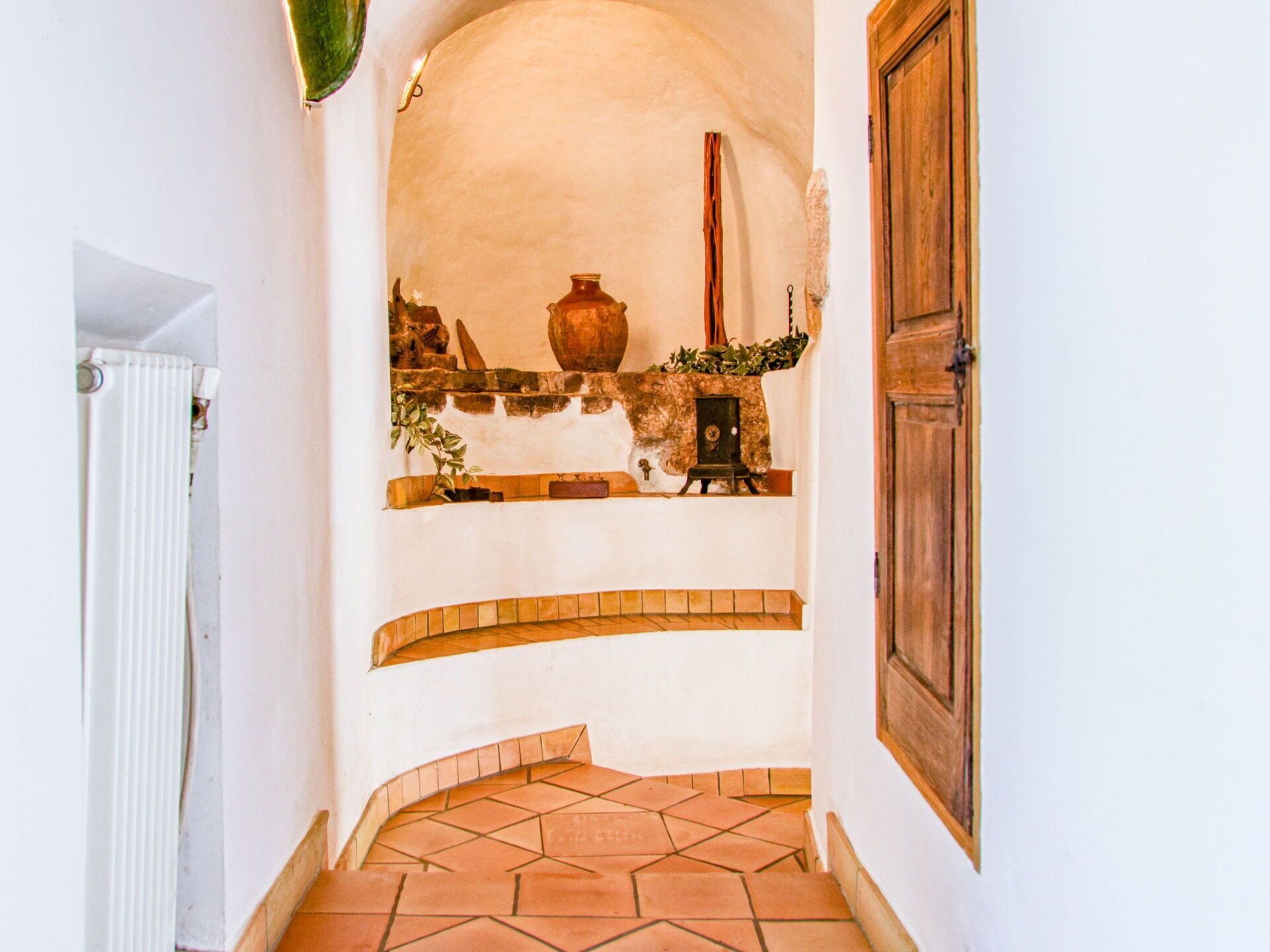 A narrow hallway with a tiled floor, a white radiator, a wooden door, and a shelf with various items including a large pot.