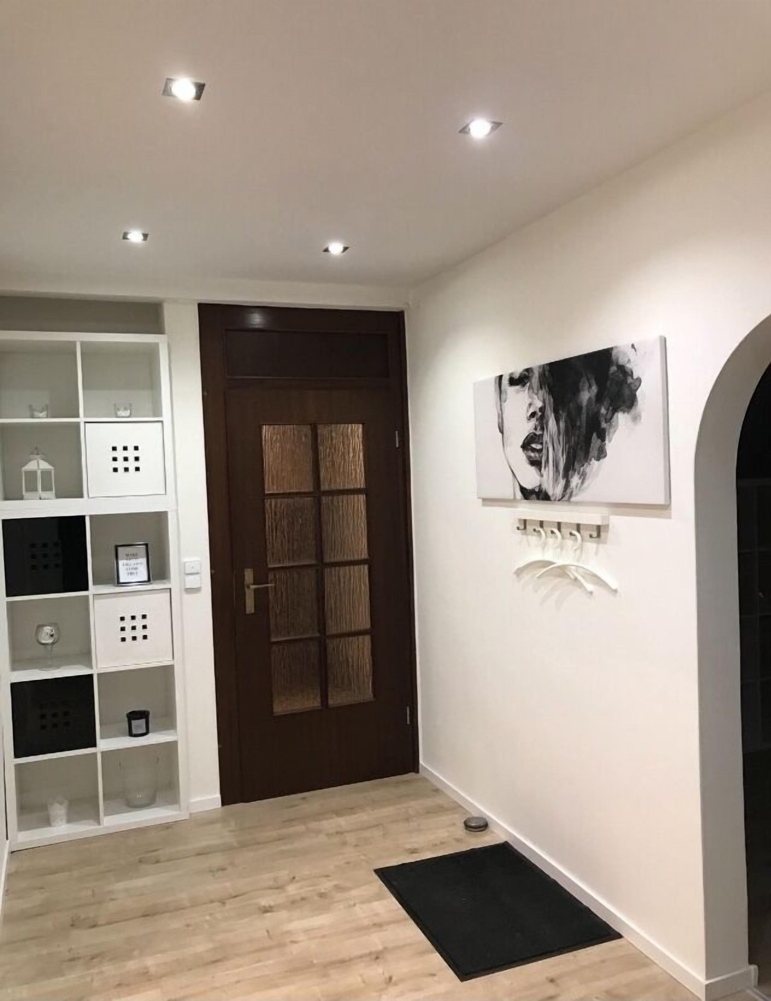 Un pasillo con una puerta de madera, un estante blanco con varios objetos y una obra de arte blanco y negro en la pared.