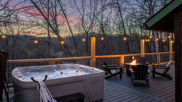 Una vasca idromassaggio su un pontile di legno, con sedie e un tavolo, circondata da alberi e luci decorative.