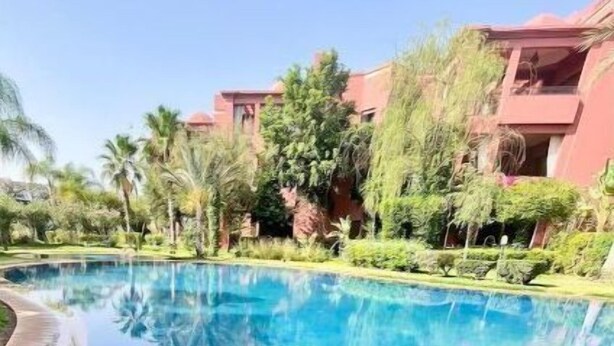 Uma piscina azul límpida com borda curva, rodeada por vegetação exuberante e um edifício com fachada vermelha.