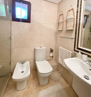 Un baño pequeño con inodoro, bidé y lavamanos. Hay un perchero sobre el lavamanos y una ventana sobre el inodoro.
