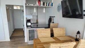 Una cocina moderna con armarios blancos, encimera negra y horno empotrado. Hay una zona de comedor con mesa de madera y sillas de mimbre.