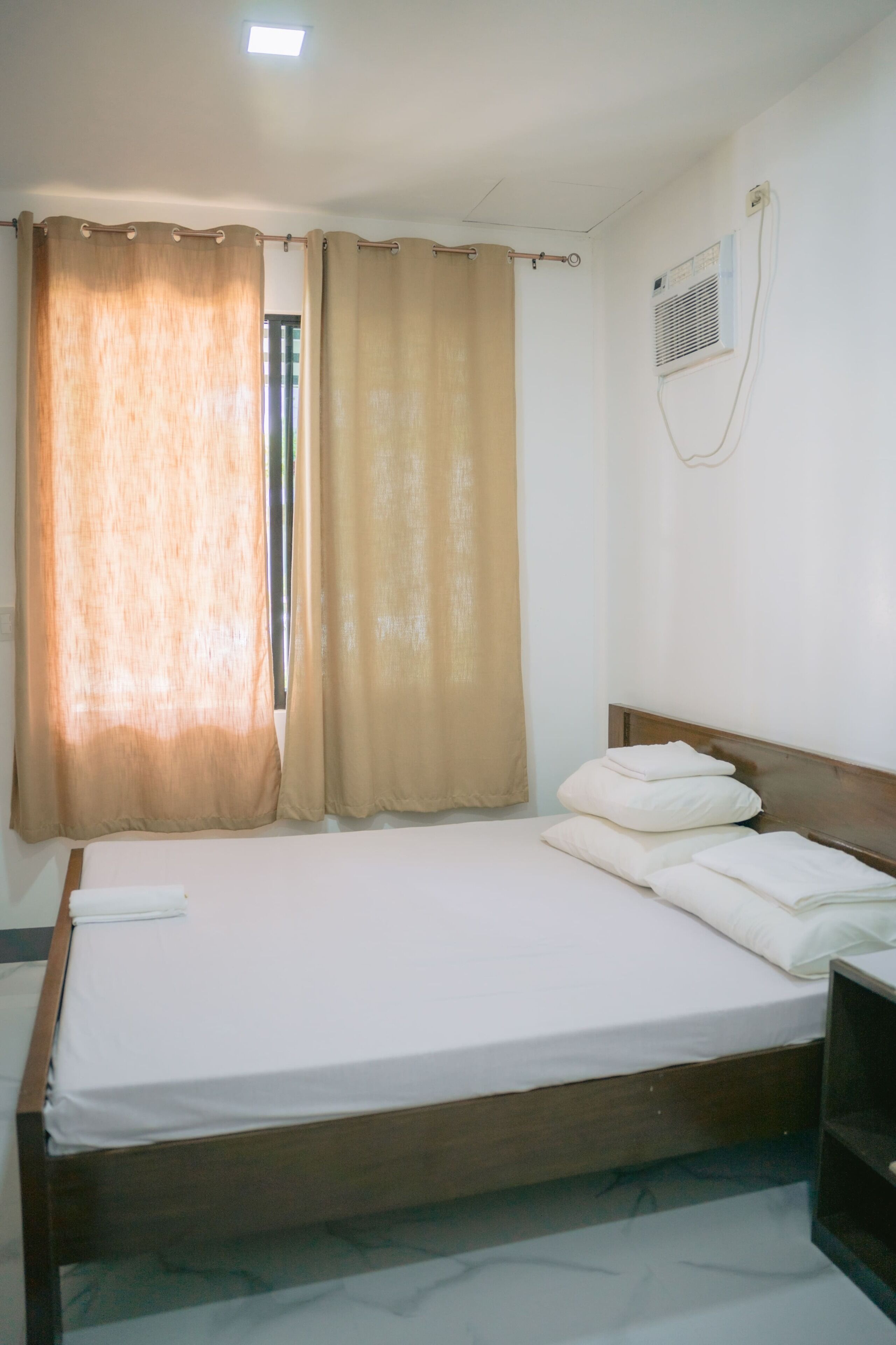 A single bed with a wooden headboard, beige curtains, and a white wall with a vent.