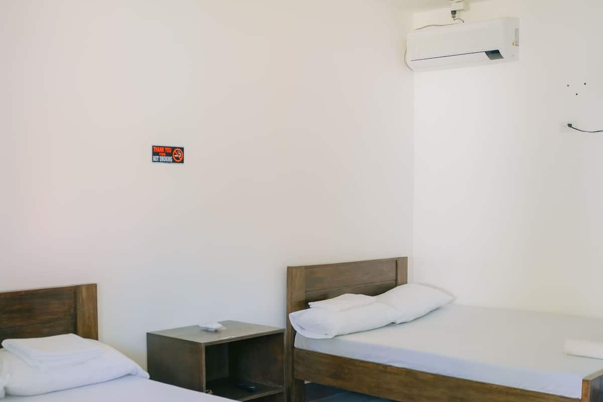 A room with two wooden beds, a small bedside table, a wall-mounted air conditioner, and a smoke detector.
