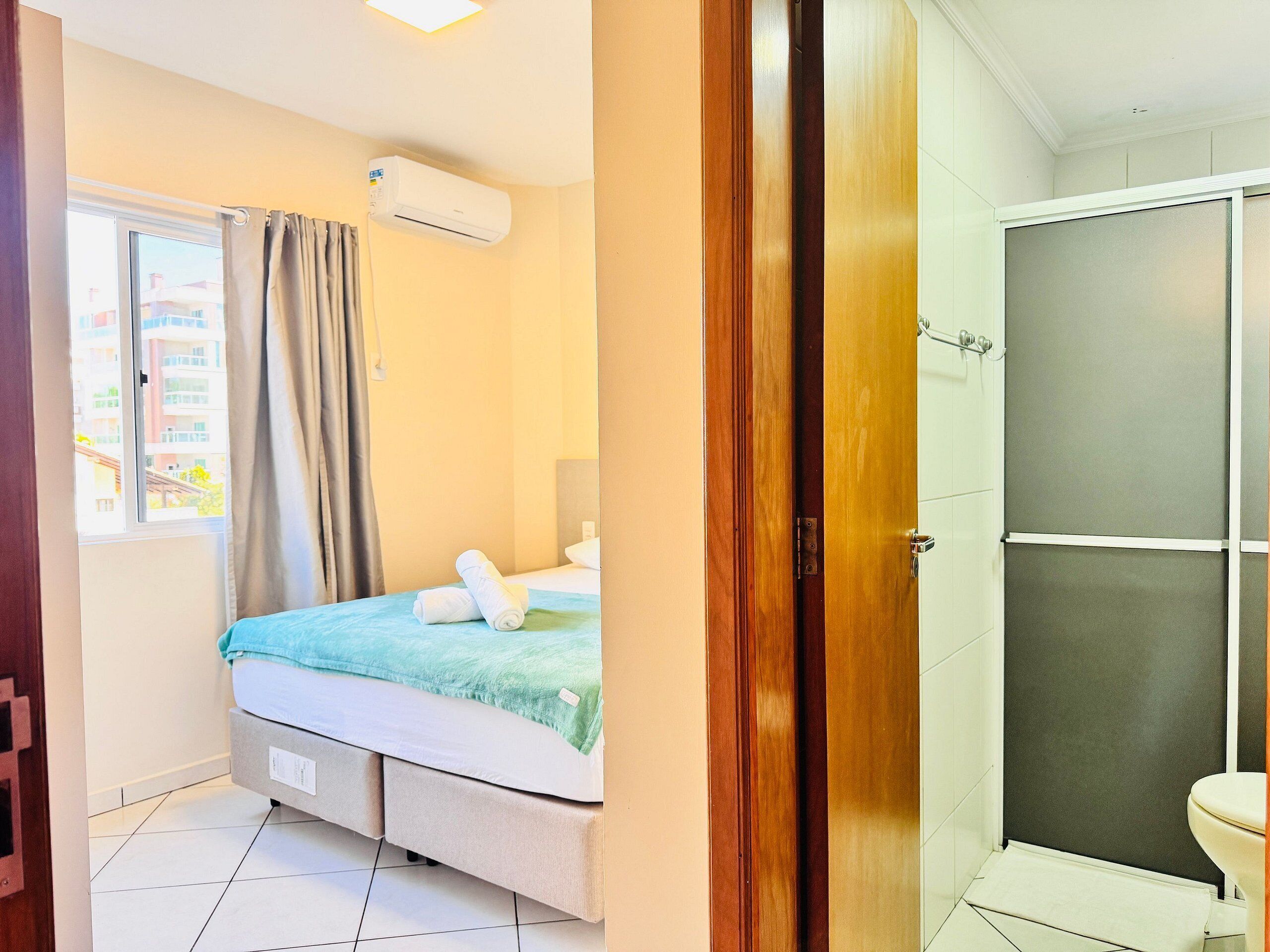 Habitación de hotel con cama, ventana con cortinas, equipo de aire acondicionado, zona de ducha y retrete.