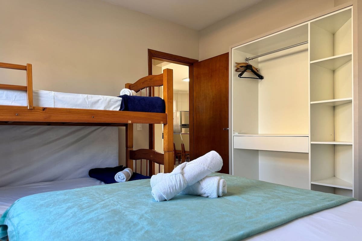 Una cama litera con estructura de madera, una cama individual con manta verde y un vestidor con estantes.
