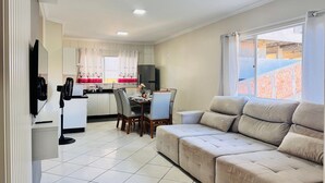 A modern living room with a beige sofa, a dining area with a table and chairs, a kitchen with a refrigerator, and a large window with a view of a building under construction. - Canto do Corrupião (Bombinhas)