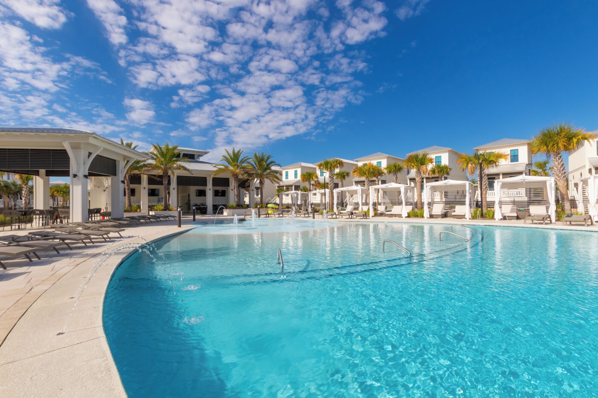 A resort pool area with a clear blue pool, lounge chairs, umbrellas, and a covered seating area.