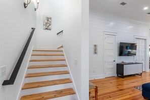 A staircase with wooden steps and a black handrail, leading to a room with white paneled walls, a flat-screen TV, and a dark-colored console table.